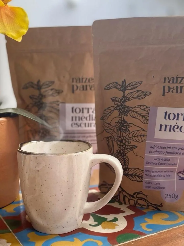 Pacotes kraft de café Raízes do Paraíso ao lado de uma caneca fumegante